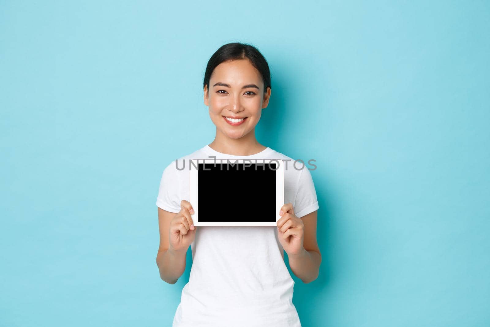 Portrait of smiling pretty asian female showing digital tablet screen with pleased expression, demonstrate shopping site, application for artist, standing light blue background.