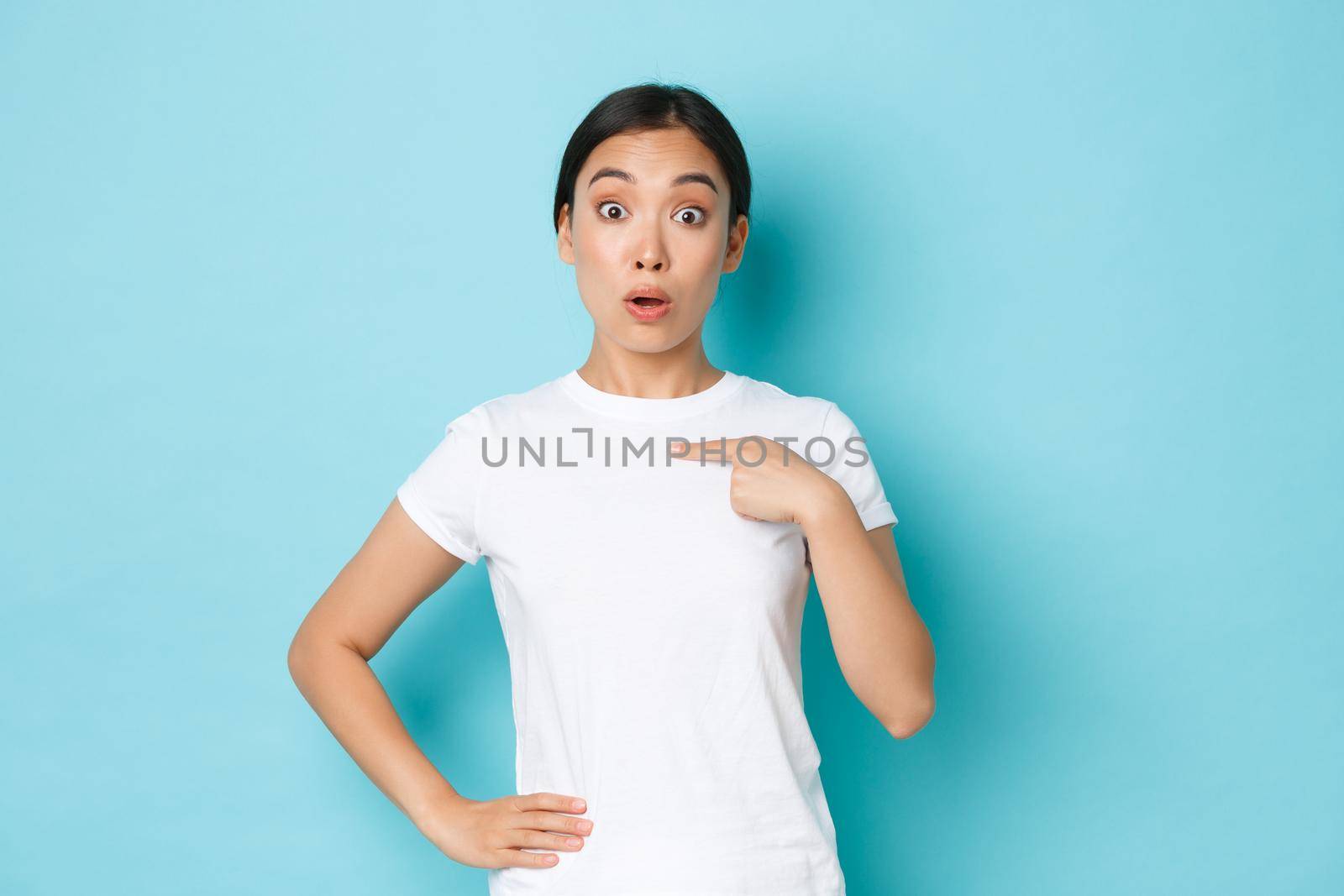 Surprised asian girl in white t-shirt pointing at herself with wondered unsure expression, being chosen or named, picked from the rest. Woman looking astonished winning something.