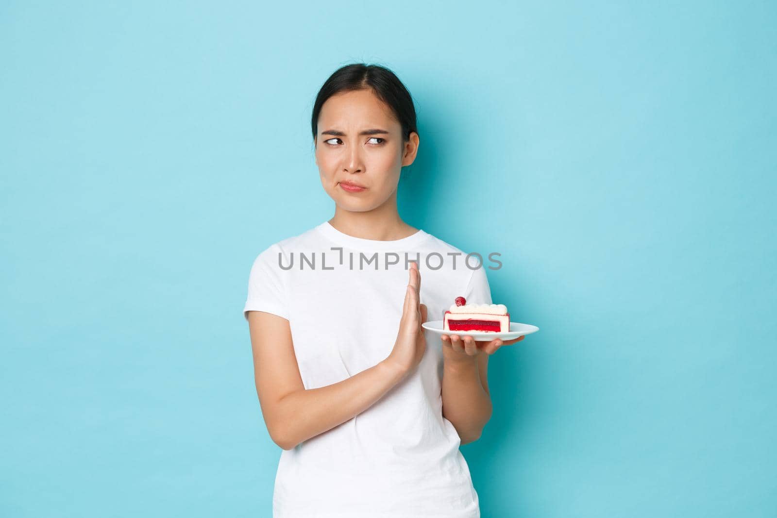 Holidays, lifestyle and celebration concept. Displeased and unamused asian female in white t-shirt, turning away from piece of delicious cake, rejecting eating dessert and grimacing.