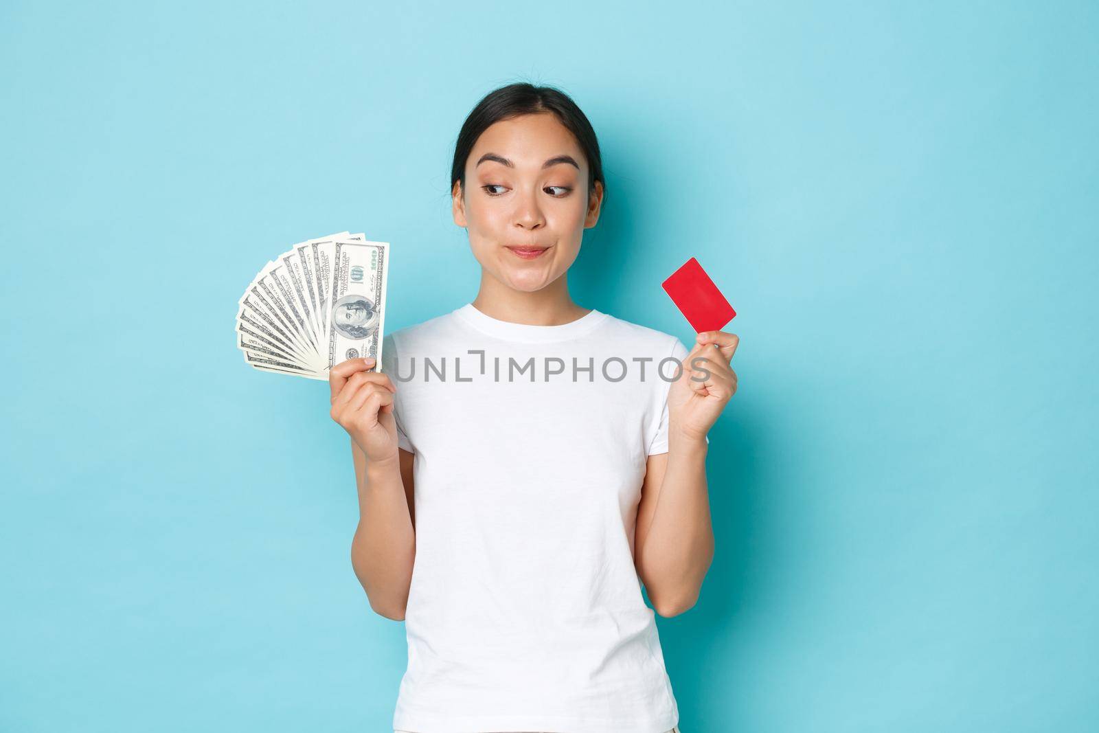 Shopping, money and finance concept. Dreamy beautiful asian girl looking happy and thoughtful upper left corner, smiling while choosing what buy on cash and with credit card by Benzoix