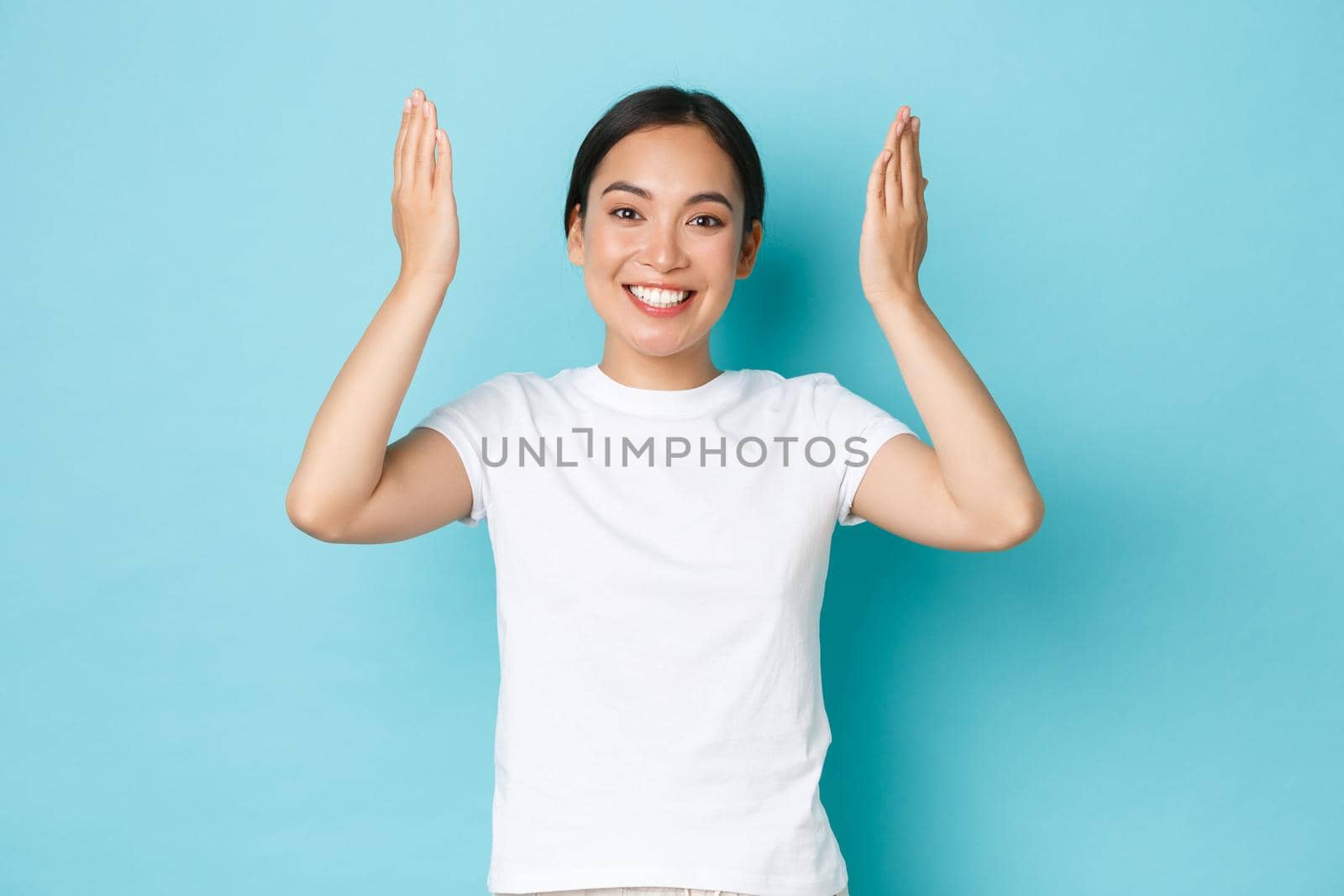 Cheerful happy asian girl open eyes and smiling cheerful and surprised, raising hands up and looking upbeat over blue background, celebrating birthday, being amazed with gift.