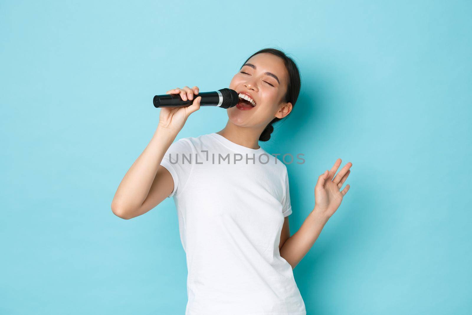 Lifestyle, people and leisure concept. Passionate and carefree pretty asian girl singing song in microphone, bending during performance, like going karaoke, standing light blue background performing.