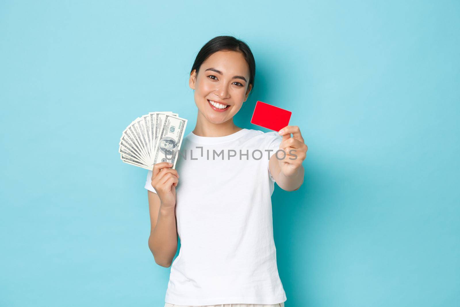 Shopping, money and finance concept. Happy carefree asian girl in white t-shirt holding cash but choosing credit card, like contactless payment, smiling upbeat, blue background by Benzoix