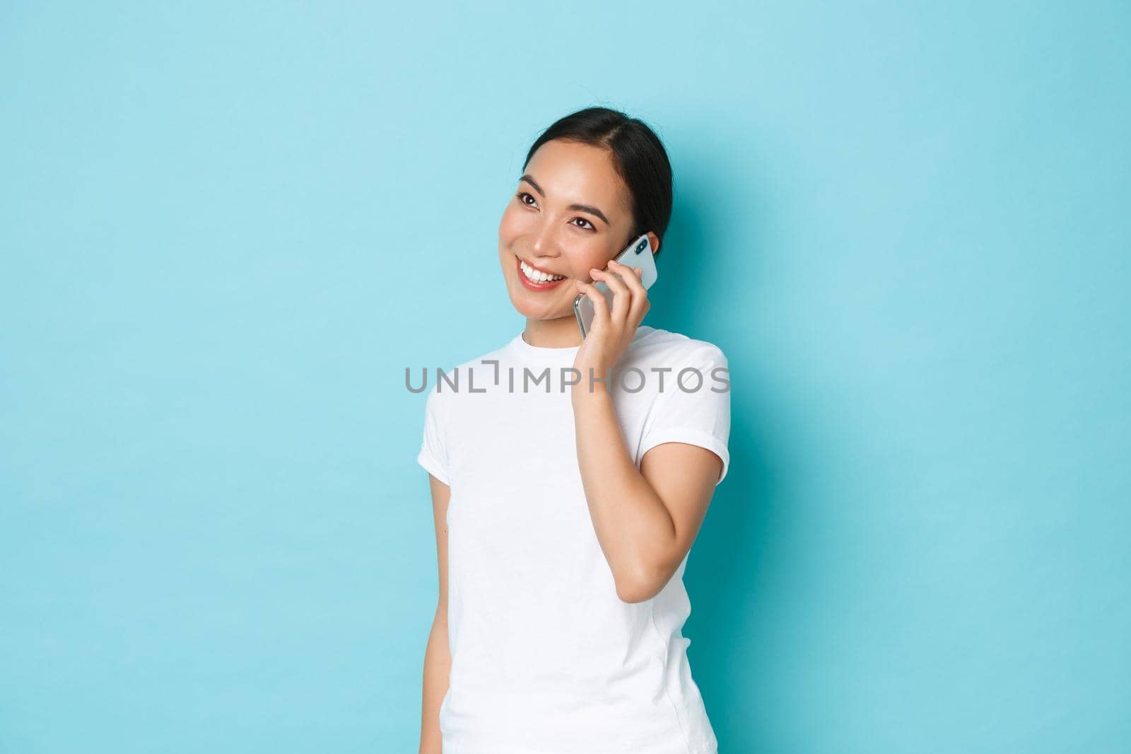 Portrait of smiling modern asian girl looking happy while talking on phone, confirm online order, chatting with friend, standing light blue background. Concept of casual conversation.