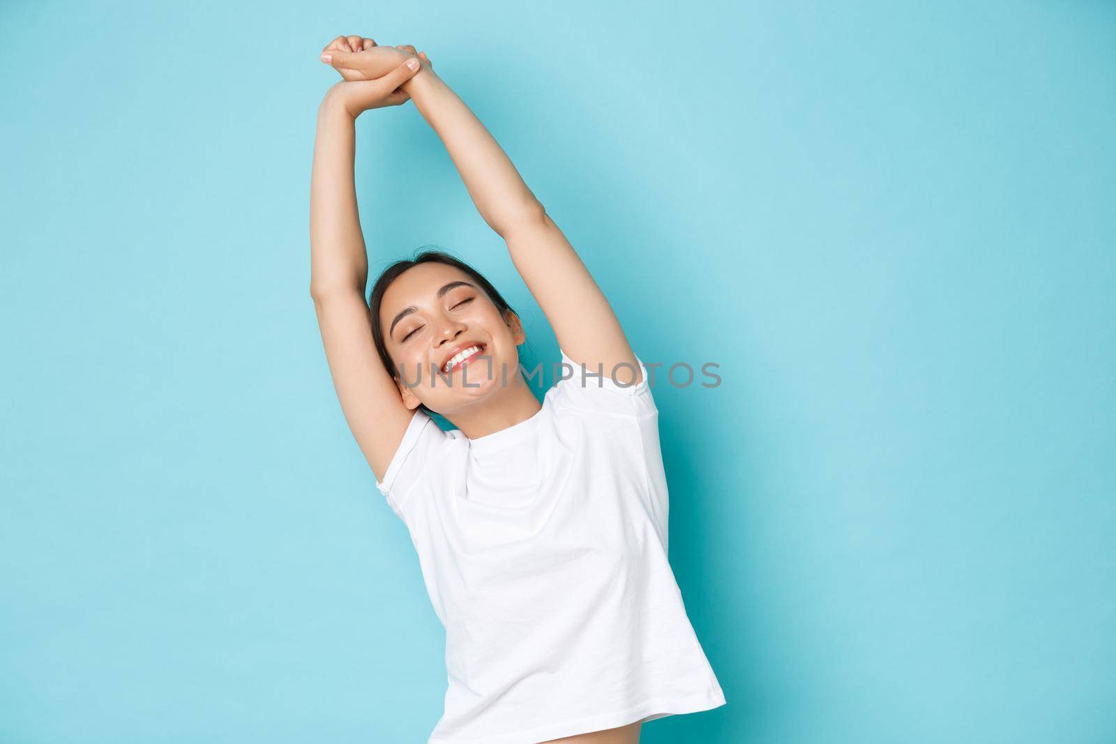 Portrait of carefree and relaxed, beautiful asian girl close eyes and stretching hands upwards with happy smile, feeling fresh and cheerful after taking nap, standing blue background.
