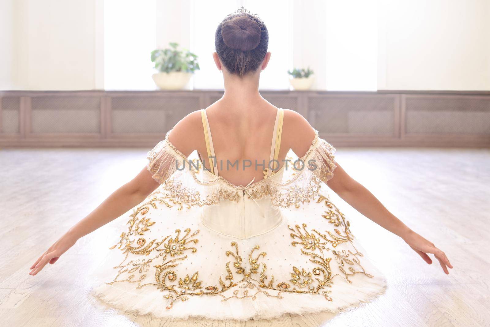 Back view of beautiful ballerina in cream dress sitting and warms up her hands on wooden floor in ballet studio by Nickstock