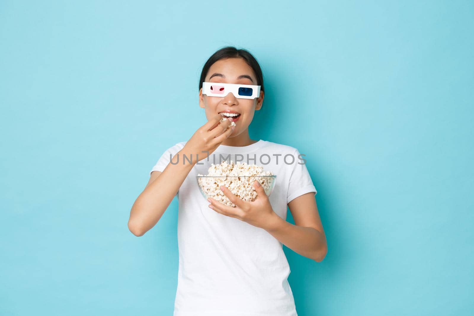 Lifestyle, leisure and emotions concept. Young excited asian girl watching premier of favorite movie, wearing 3d glasses and eating popcorn with thrilled expression, standing light blue background.