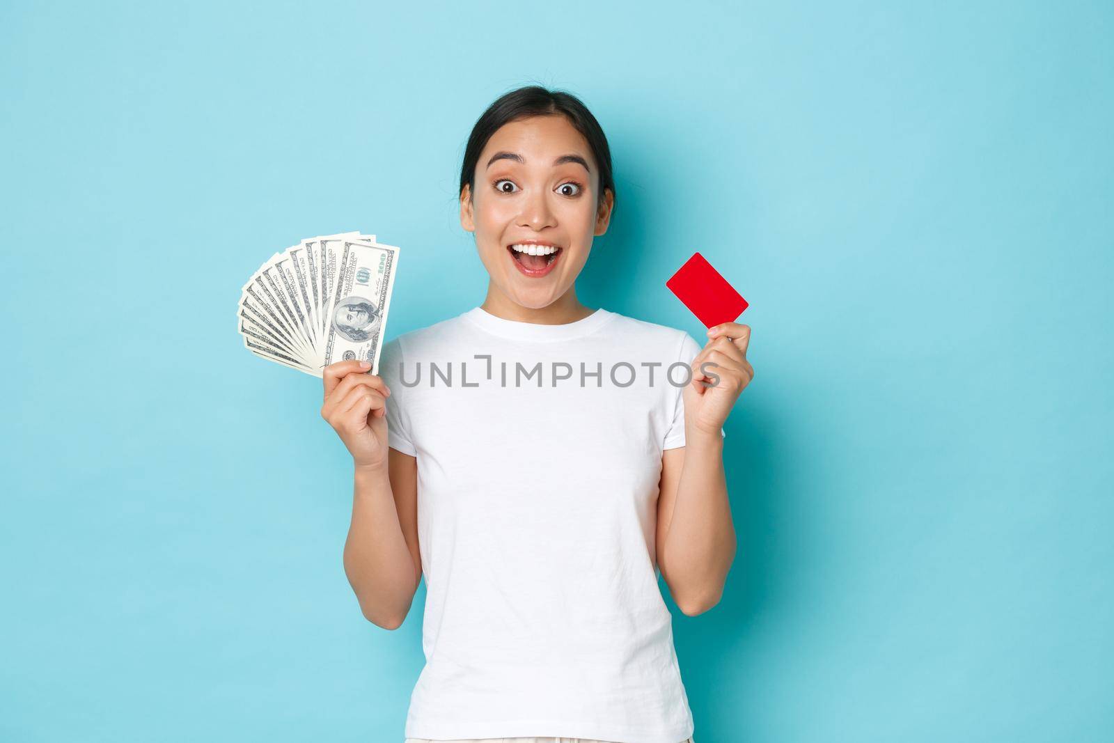 Shopping, money and finance concept. Amused asian girl in white casual t-shirt gasping, found out awesome prices, discount offers in shop, holding both credit card and cash, light-blue background by Benzoix