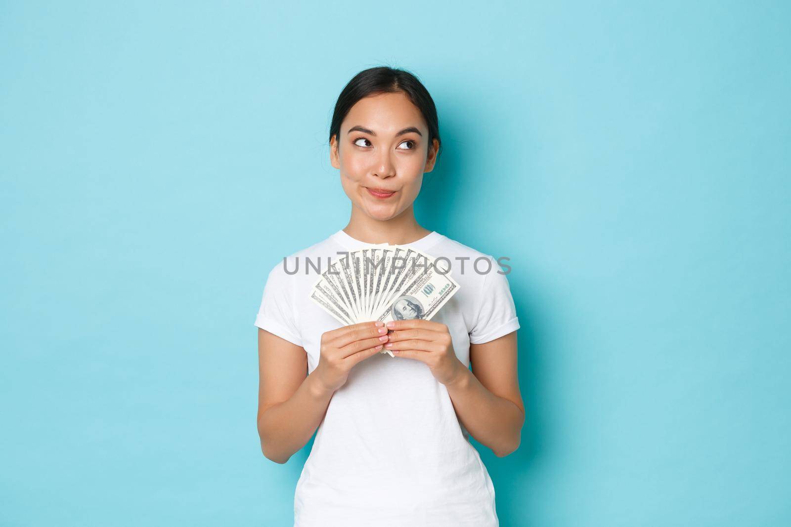 Shopping, money and finance concept. Thoughtful and creative pretty asian girl thinking where invest money, holding cash and looking upper right corner, making choice, deciding what buy by Benzoix