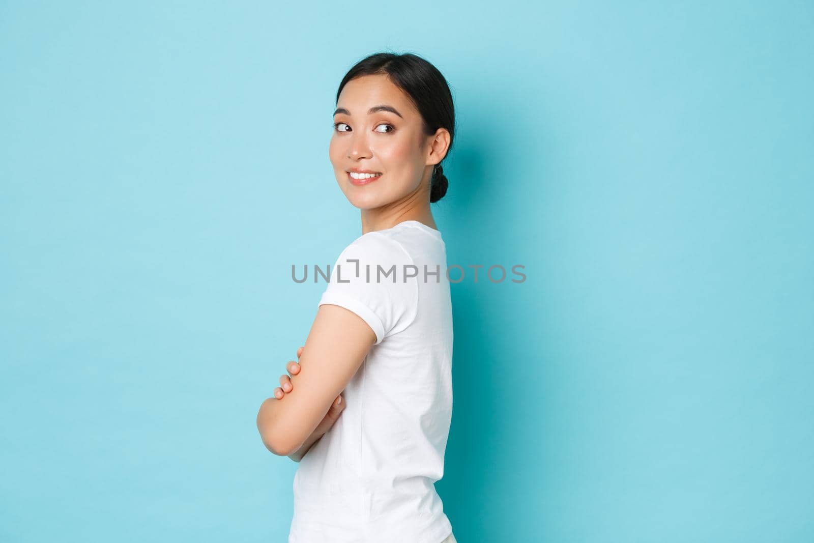 Coquettish pretty asian girl in white t-shirt standing in profile and turning right with curious smiling face, looking at something interesting, found good promo offer, standing blue background.