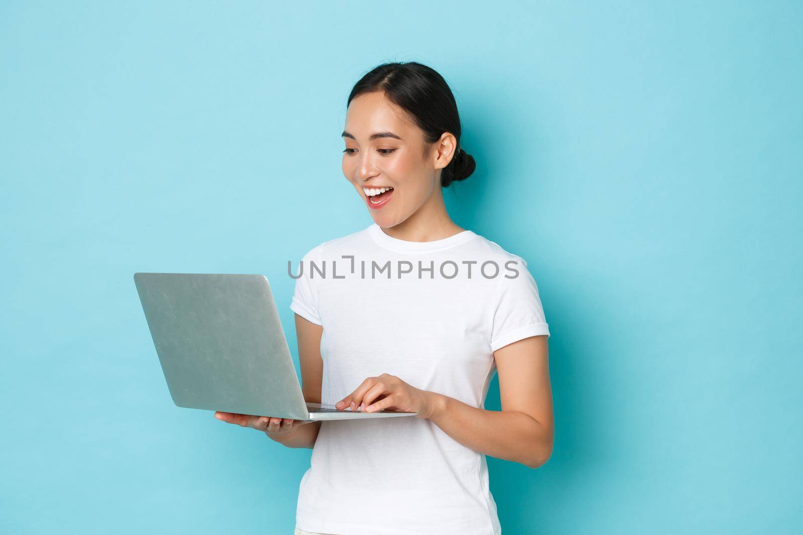 Smiling pretty asian girl in white t-shirt, working on project, looking satisfied and happy at laptop screen, freelancing or browing internet, online shopping over light blue background.