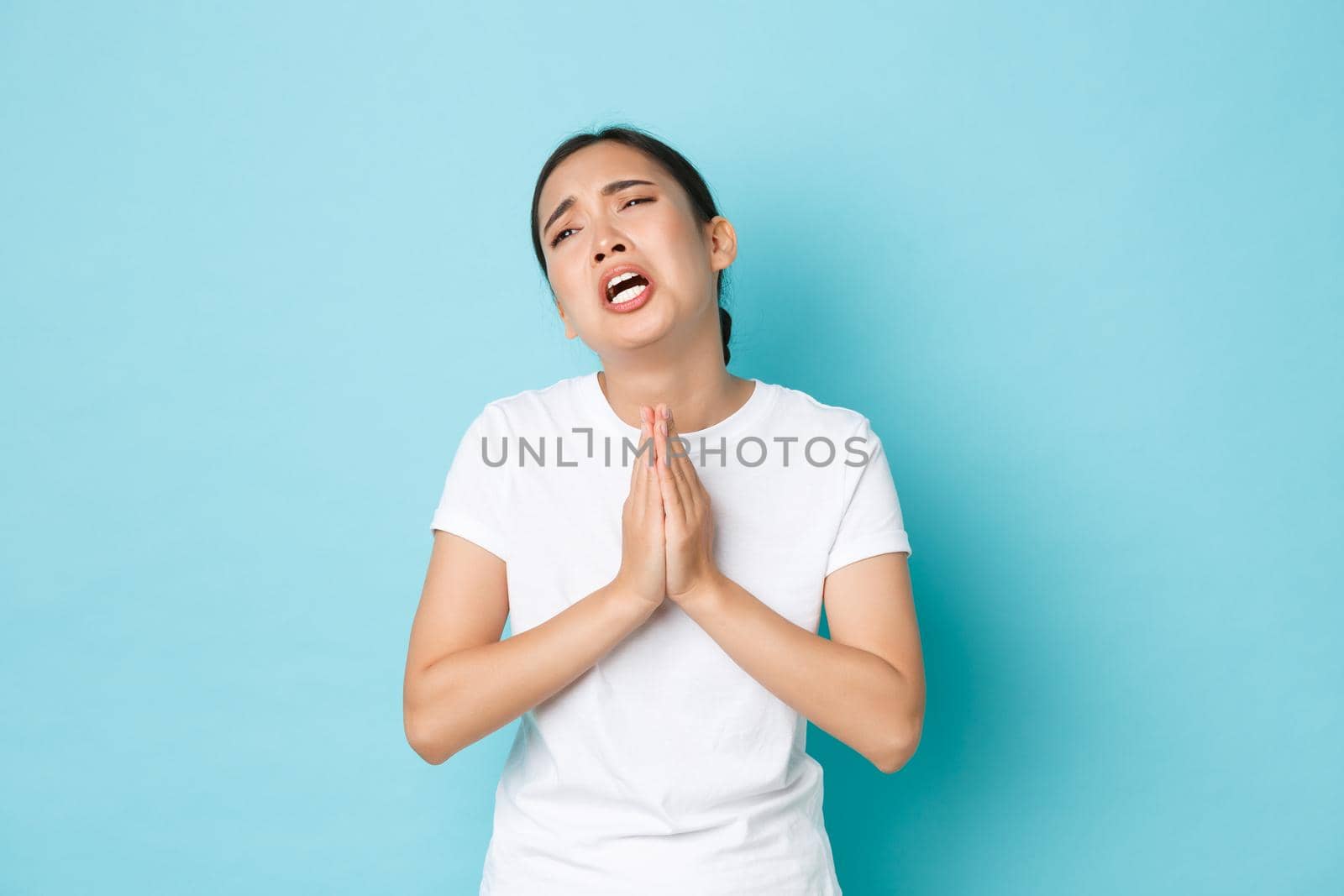 Desperate uneasy sad asian girl pleading for help, begging and crying, supplicating from troublesome life, standing blue background in need, grieving or asking for apology.