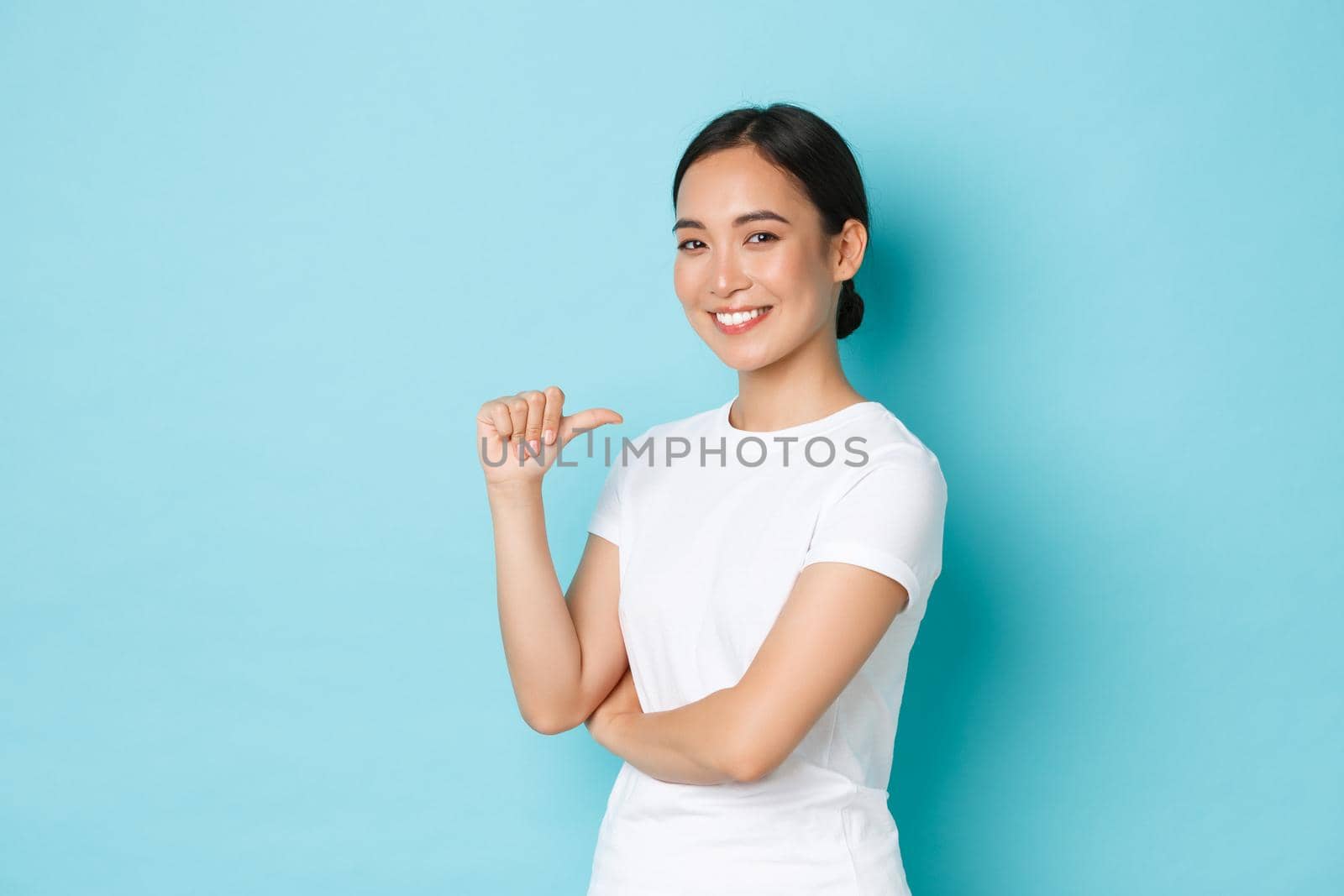 Confident smiling asian girl in white t-shirt pointing at herself with proud, assertive expression, show-off, promote own abilities, being professional, searching for job, standing blue background.