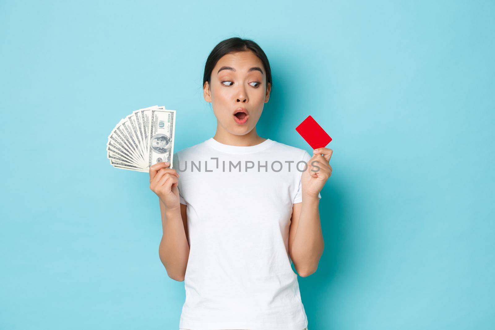 Shopping, money and finance concept. Amazed beautiful asian girl in white t-shirt gasping astonished and looking at credit card while holding cash in other hand, prefer contactless payment.