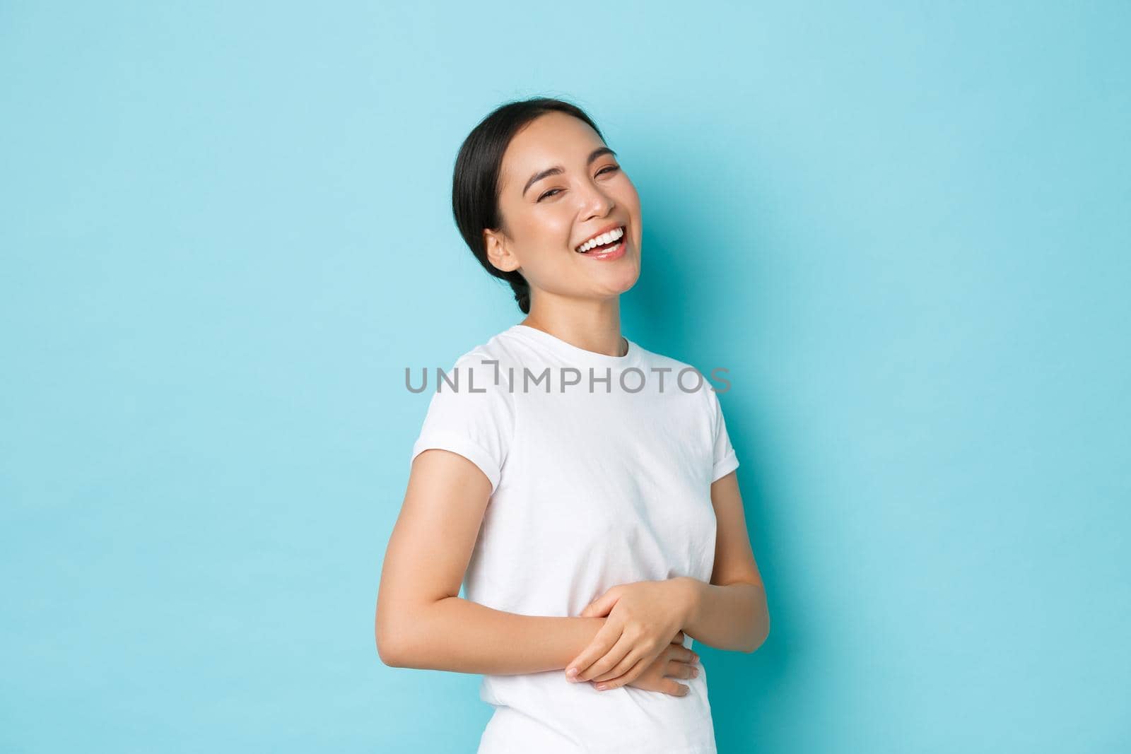 Portrait of carefree beautiful asian girl laughing and smiling pleased, holding hands on belly, standing in determined and confident pose over blue background. Copy space