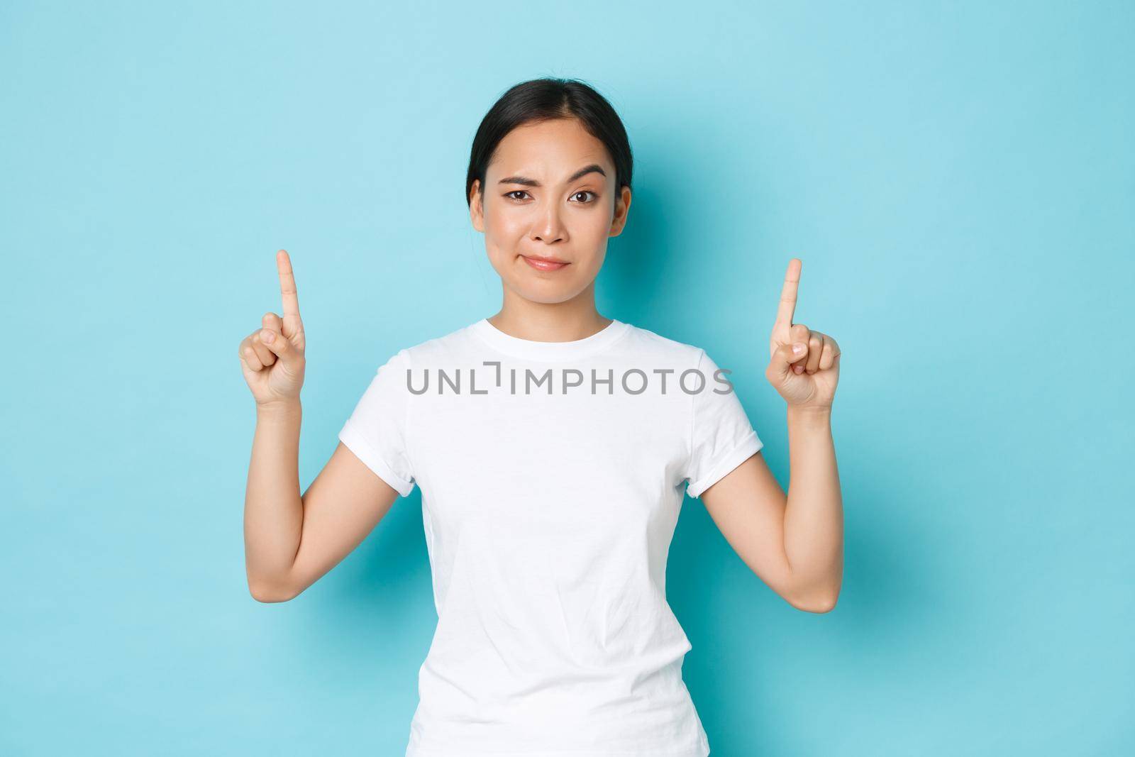 Skeptical and suspicious asian female in white t-shirt, frowning dubious, pointing fingers up at empty space, waiting for explanation, standing bothered over blue background. Copy space