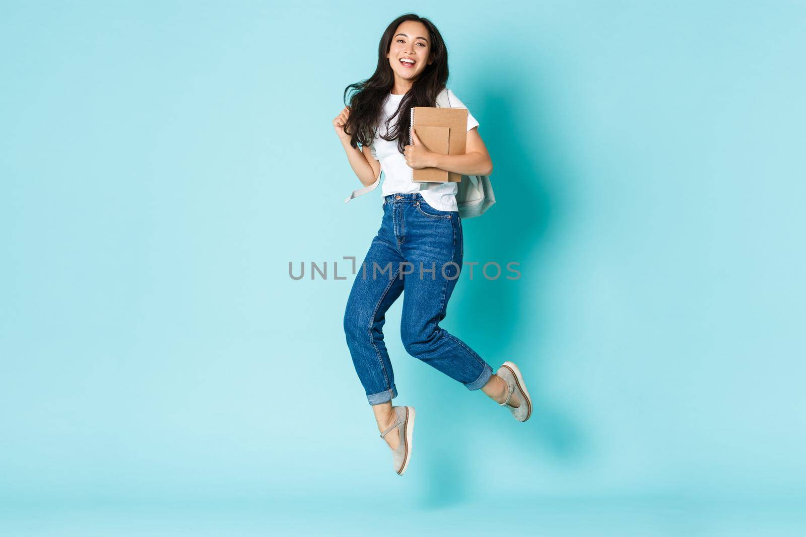 Fashion, back to school and lifestyle concept. Cheerful young asian girl, korean student looking upbeat, holding backpack and notebooks, jumping upbeat over light blue background.