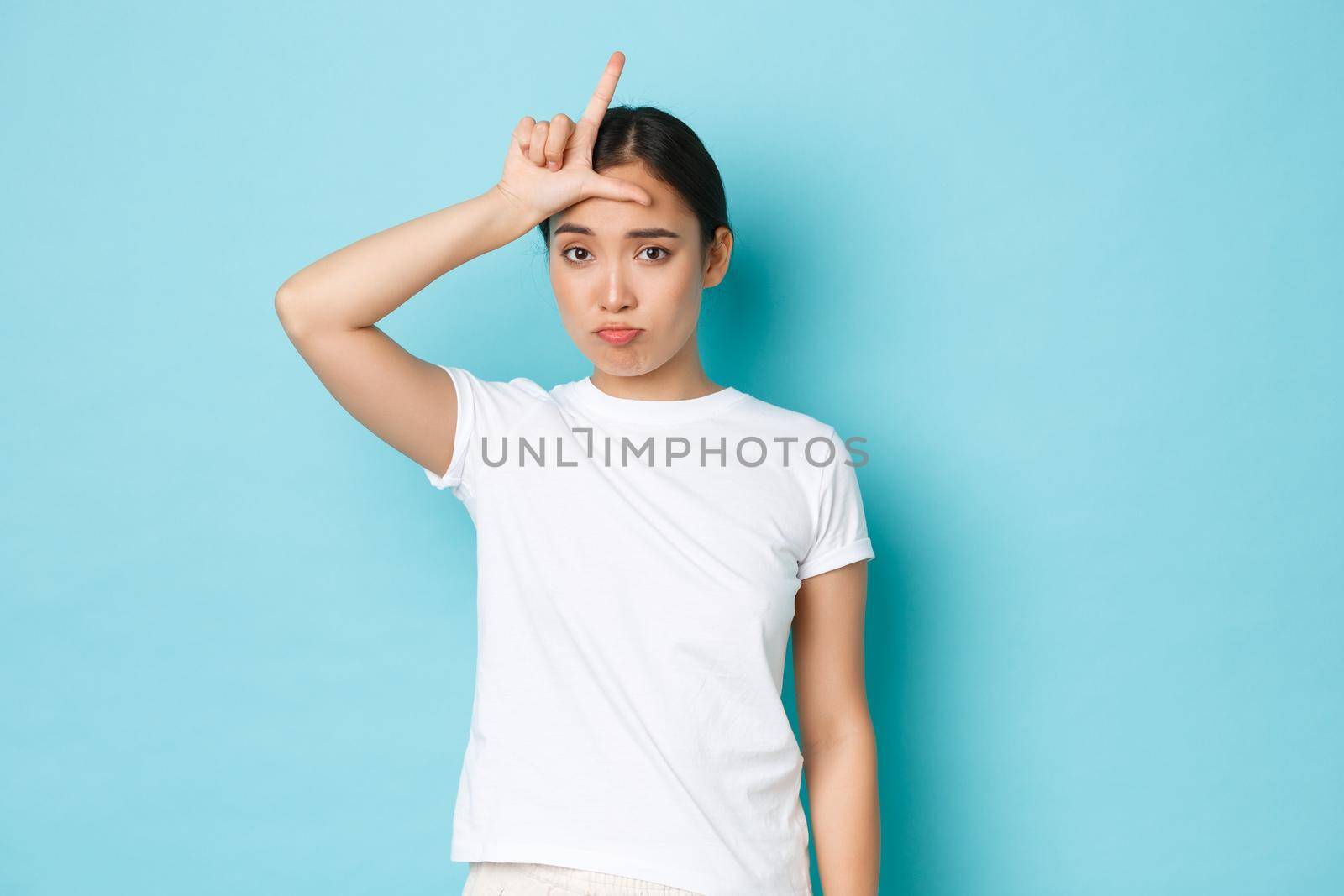 Sad and unconfident in herself asian girl wearing white casual t-shirt, sulking and looking glomy over lost competition, showing loser gesture on forehead, standing blue background.