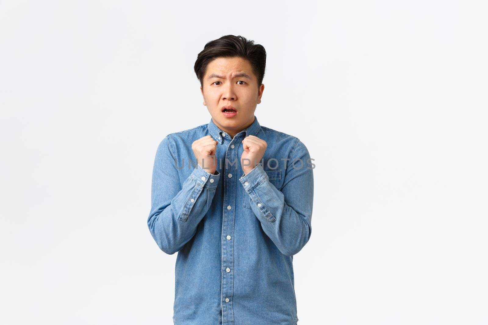 Portrait of timid and insecure young asian man feeling cornered or scared, holding hands tight to chest, shivering fear, looking anxious at camera, standing frightened over white background.