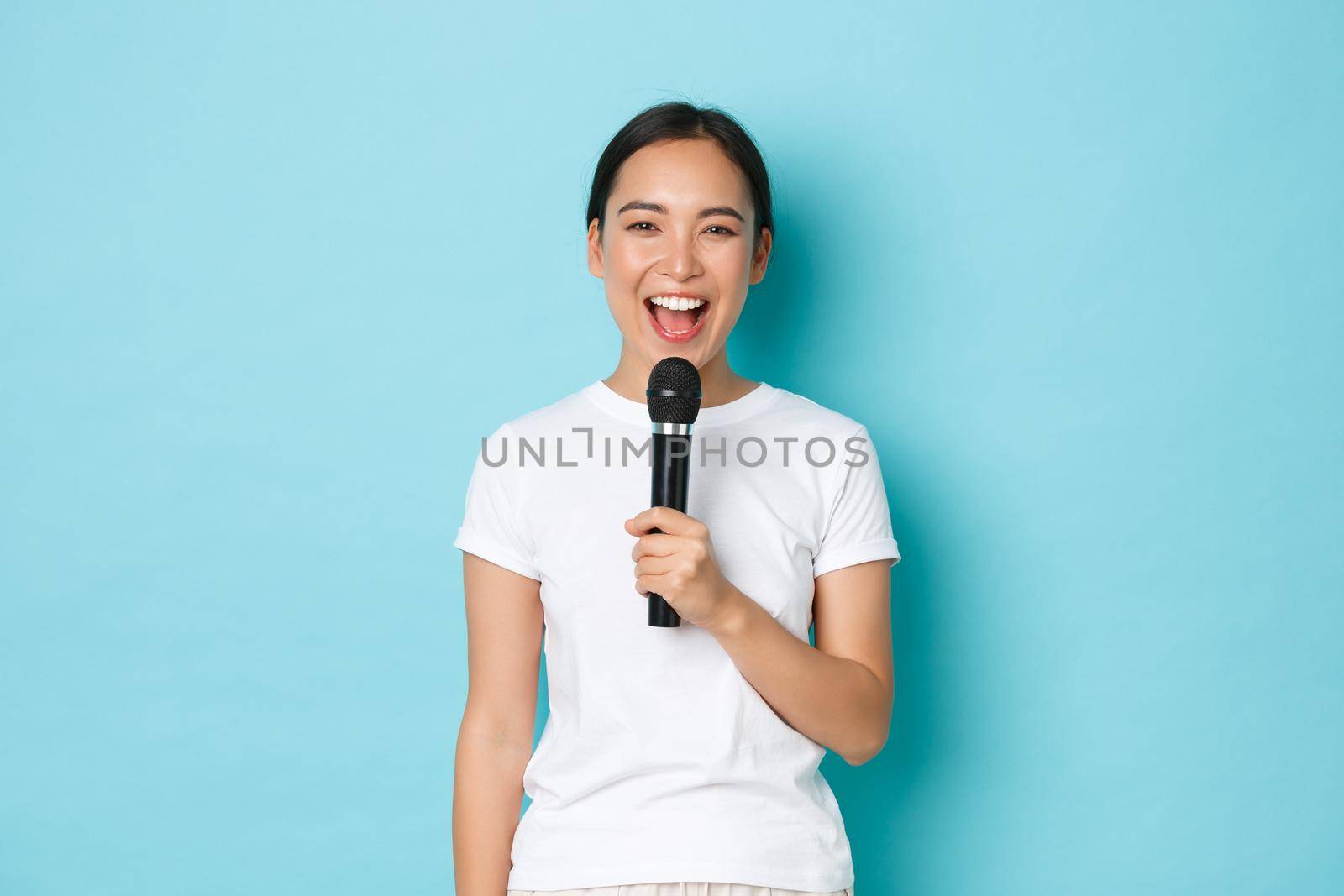 Lifestyle, people and leisure concept. Happy carefree asian girl enjoying performing song, holding karaoke microphone and smiling, singing favorite song, standing blue background by Benzoix