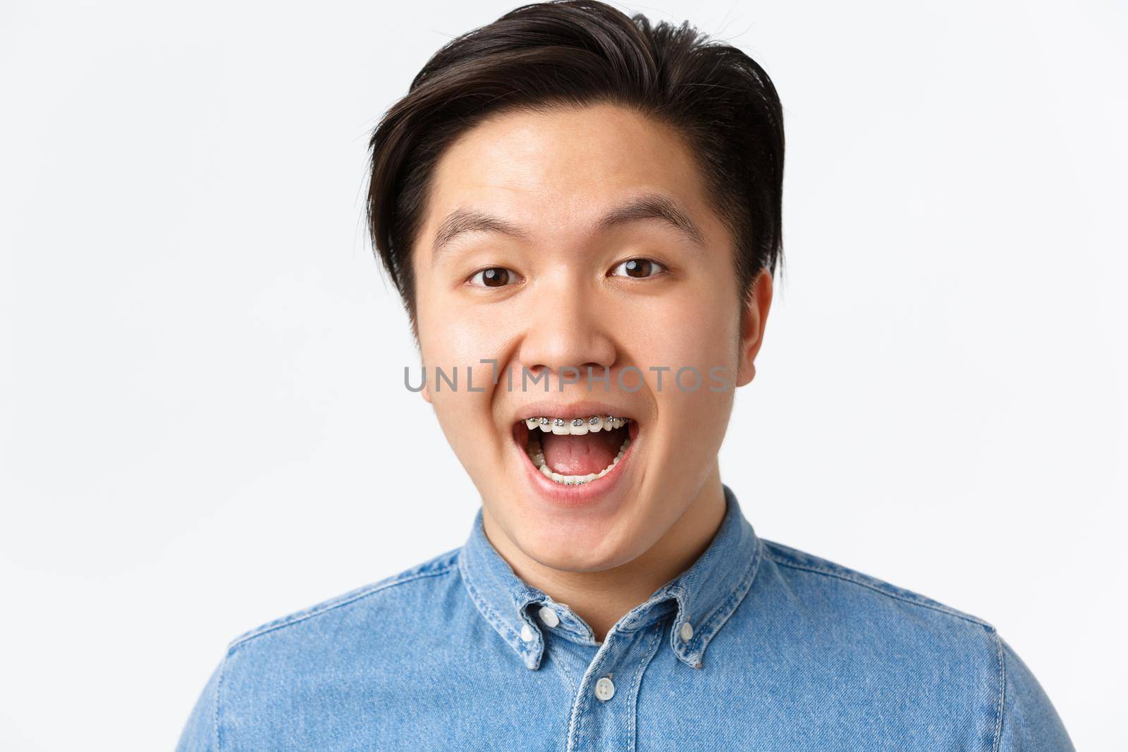 Orthodontics, dental care and stomatology concept. Close-up of surprised enthusiastic asian man looking amused and smiling, showing teeth braces, standing white background.