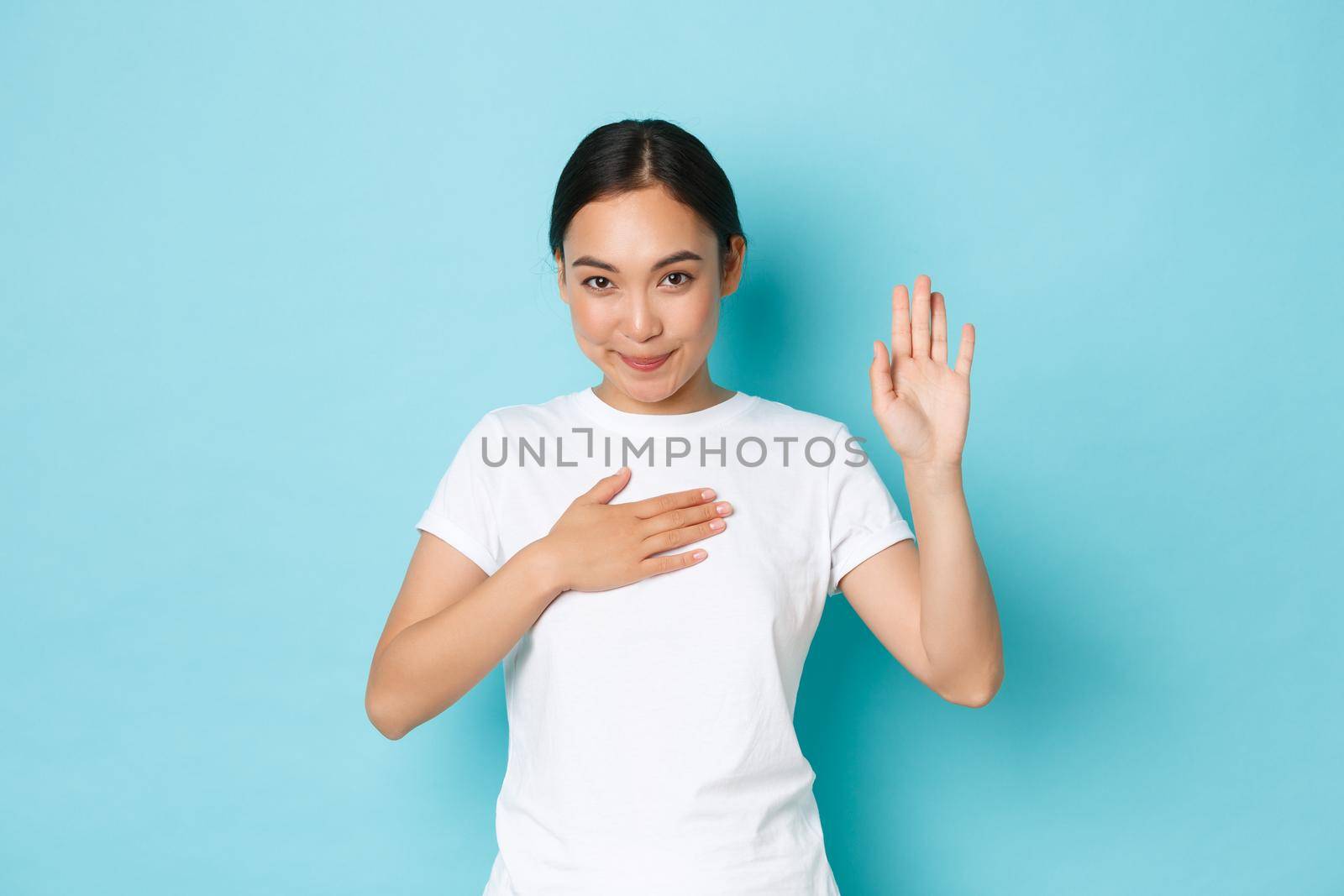 Cute smiling asian girl swearing to tell only truth, making oath or pledge on something. Adorable korean female student hold hand on heart and arm raised while being honest, blue background.
