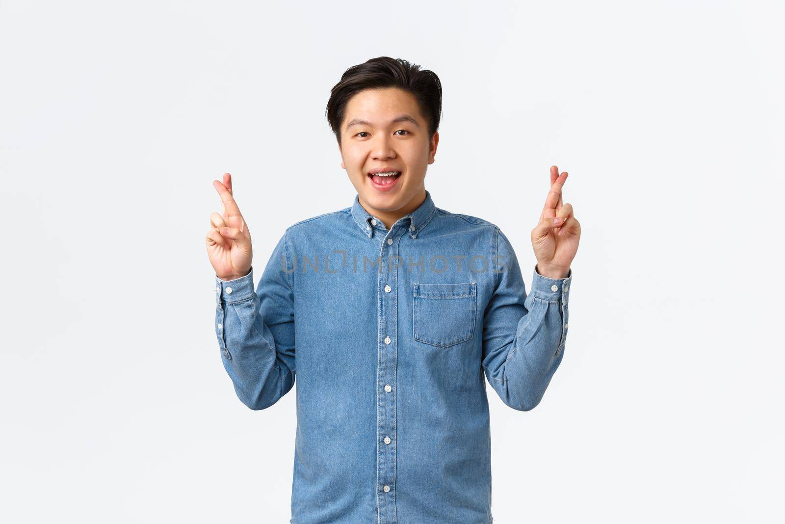 Portrait of hopeful optimistic asian man with braces, smiling upbeat, having faith in dreams come true. Guy making wish with fingers crossed, anticipating miracle, feeling lucky, white background.