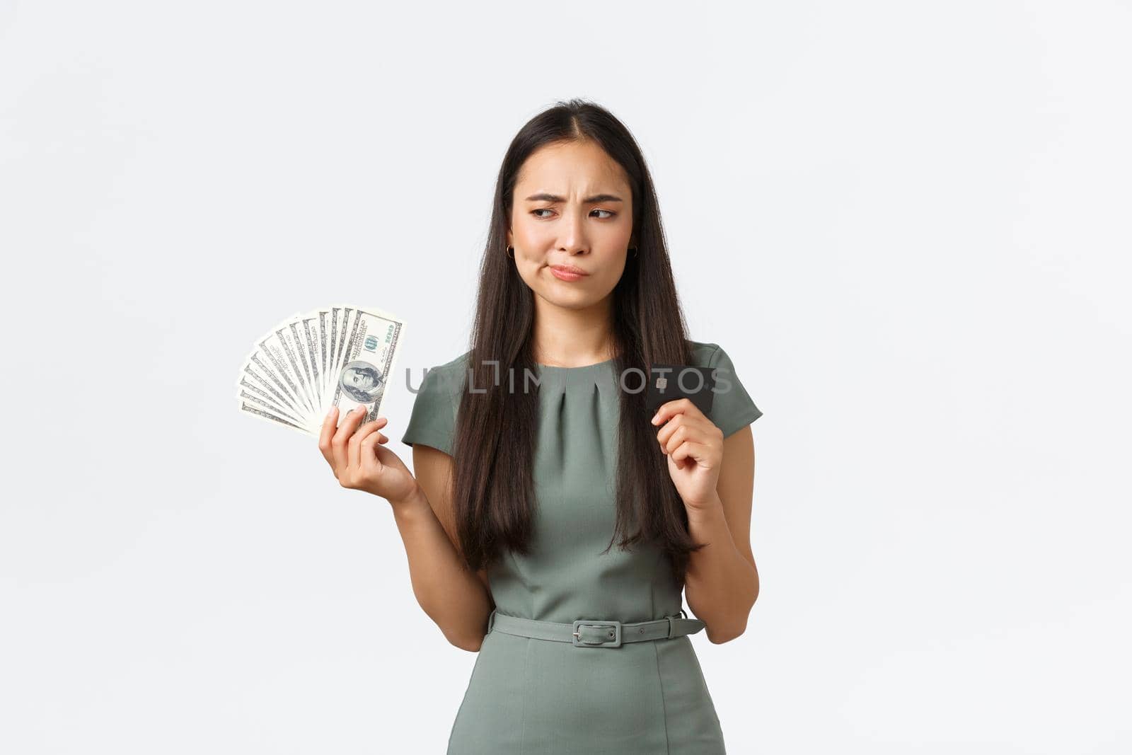 Small business owners, women entrepreneurs concept. Indecisive asian woman thinking while holding credit card and cash, perplexed how to pay for purchase, standing white background thoughtful by Benzoix