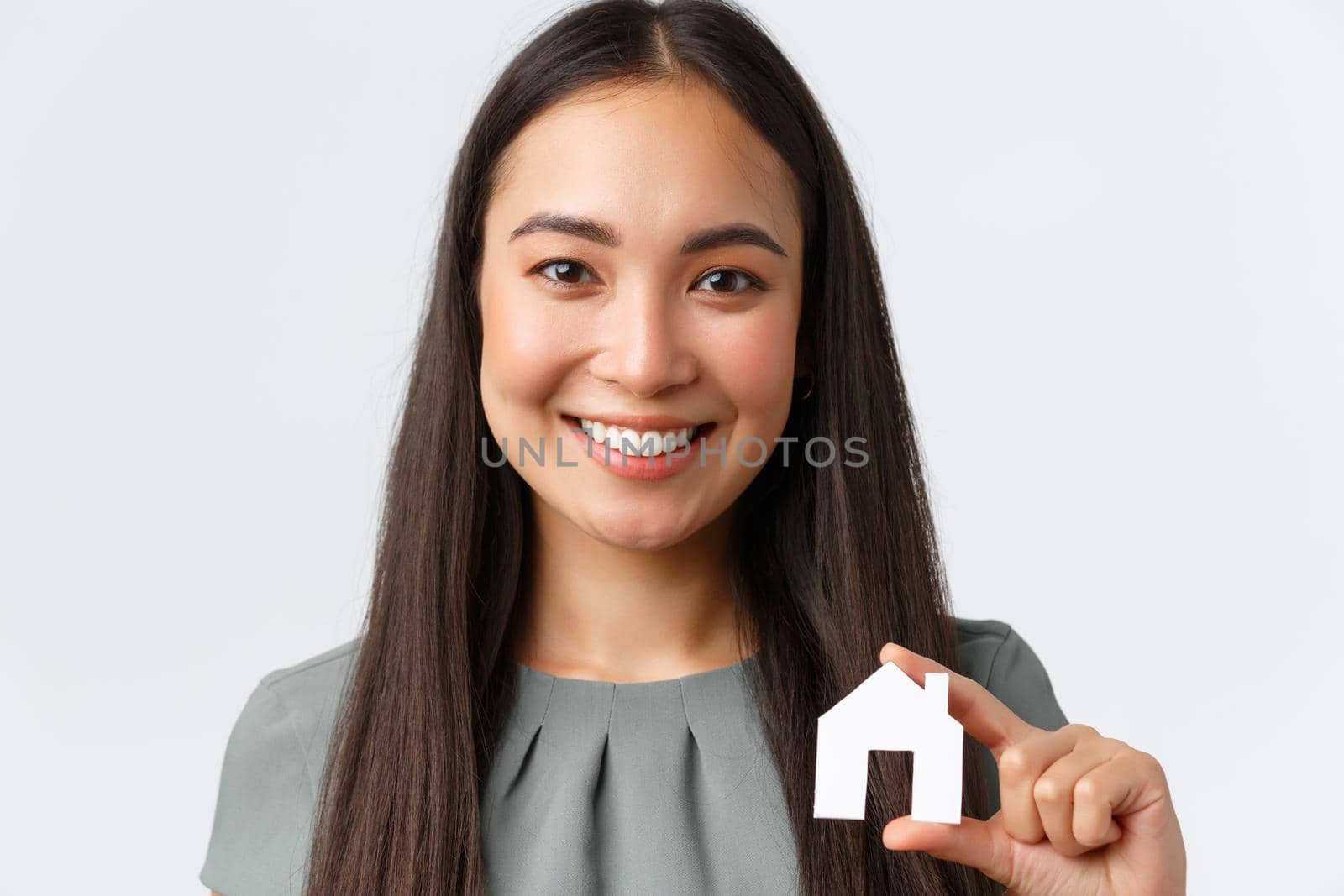 Insurance, loan, real estate and family concept. Close-up of happy smiling pretty asian woman showing small miniature of home, holding paper house, selling or renting apartment, white background.