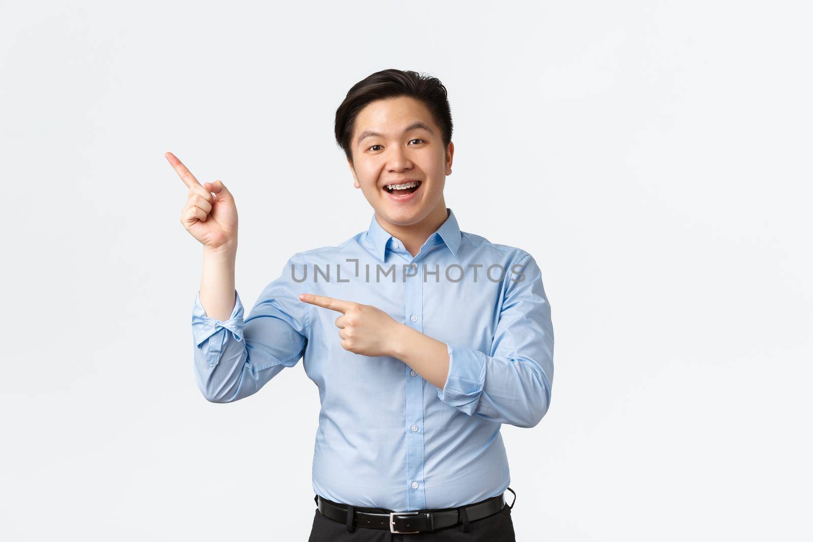 Business, finance and people concept. Enthusiastic smiling asian male entrepreneur in blue shirt, with teeth braces, pointing fingers upper left corner, making announcement, white background.