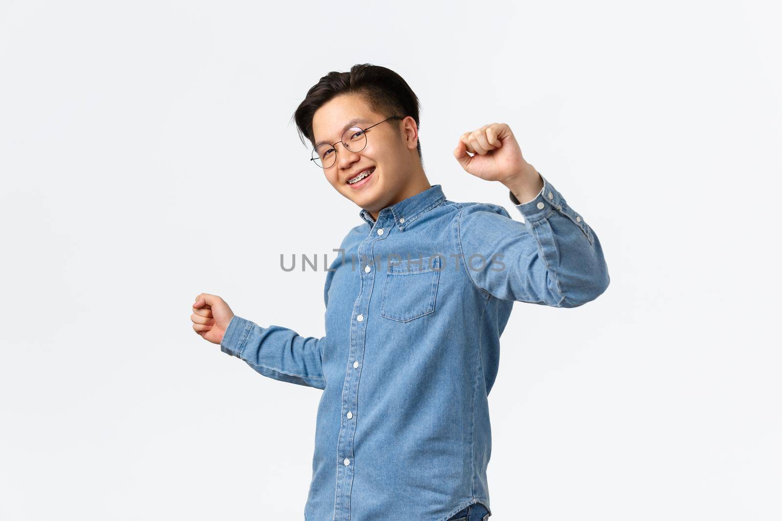 Lifestyle, leisure and technology concept. Young carefree asian man looking upbeat and unbothered, wearing glasses, put braces in dental clinic, dancing happy over white background, smiling.