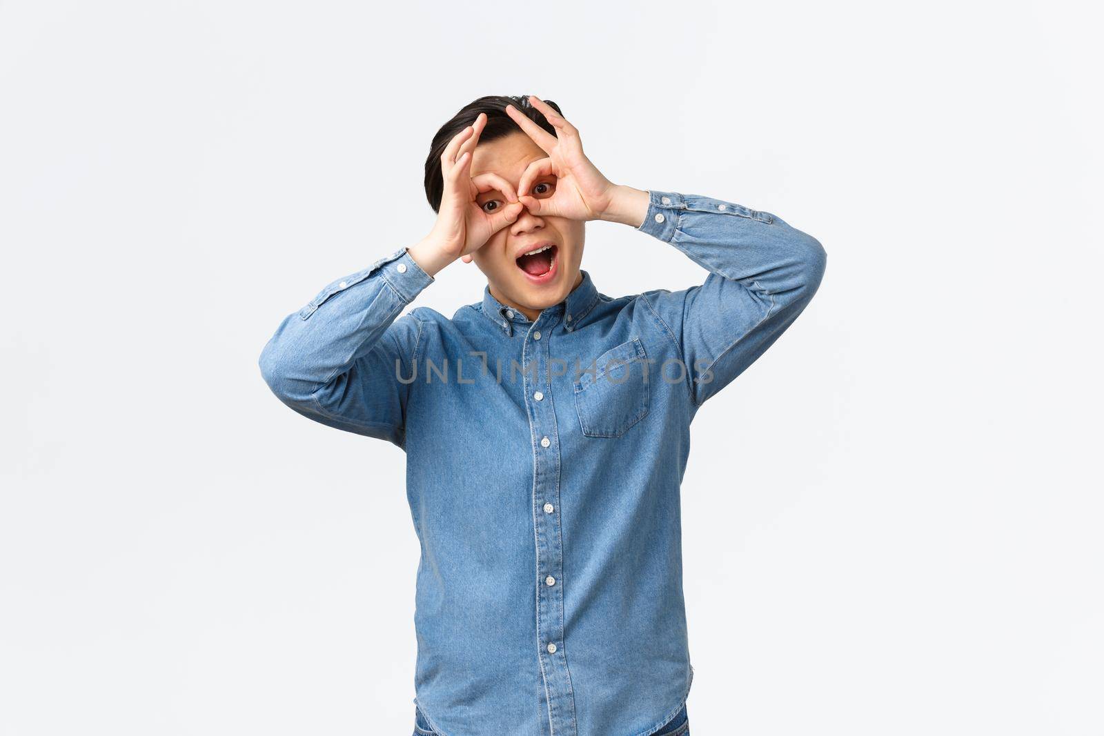 Funny and upbeat, playful asian man making faces, showing fake glasses with hands over eyes, mocking someone, playing, seeing something exciting, standing white background.