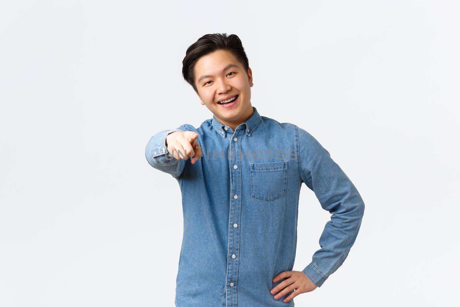 Smiling happy asian handsome guy in blue shirt, have braces, pointing finger at camera, encourage person click link, naming or choosing someone, standing over white background by Benzoix