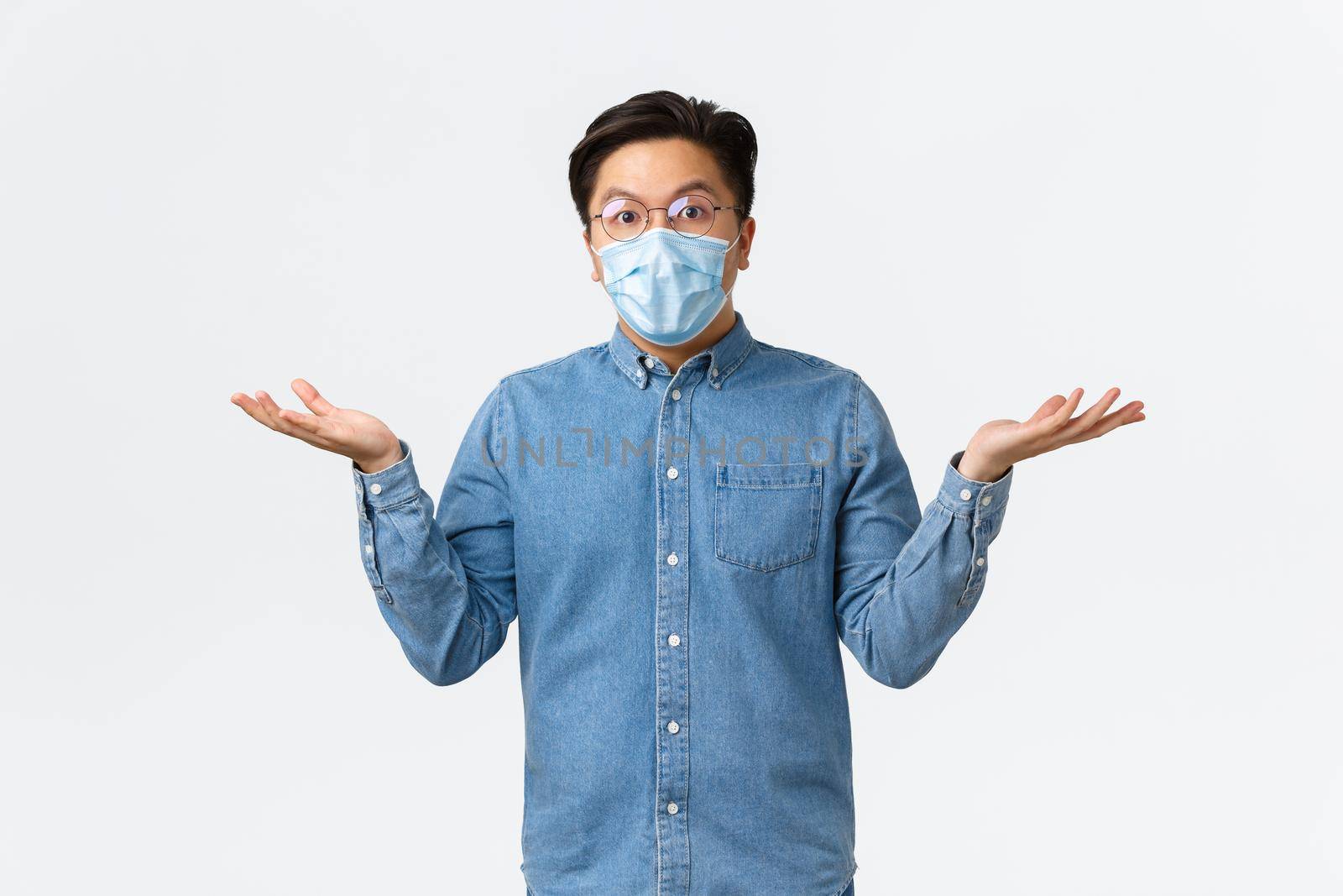 Covid-19, preventing virus, and social distancing at workplace concept. Confused and surprised asian male entrepreneur spread hands sideways and shrugging puzzled, wear medical mask and glasses by Benzoix