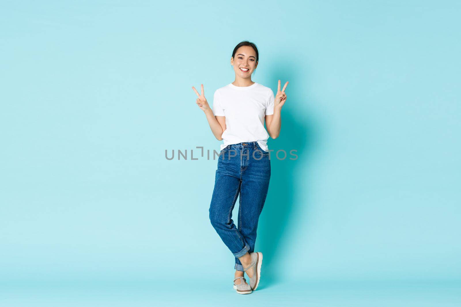 Fashion, beauty and lifestyle concept. Full-length portrait of adorable smiling asian woman in casual outfit, showing peace gestures and looking happy, standing light blue background.