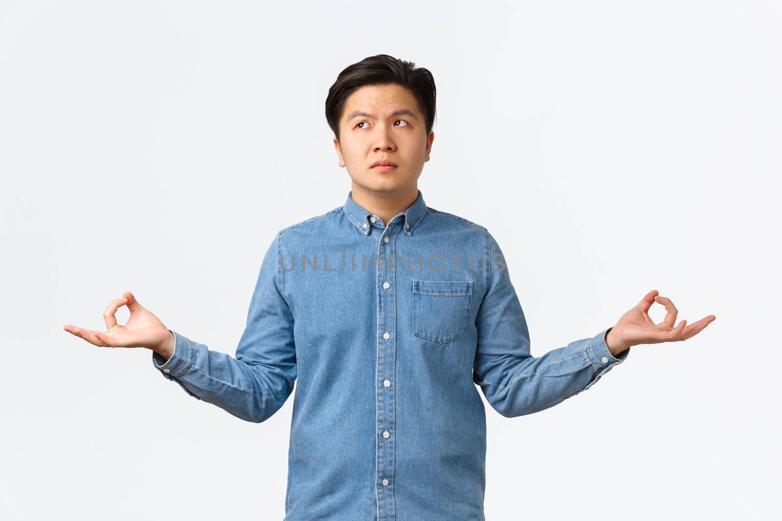 Uneasy and concerned asian man looking away thoughtful, overthinking as trying to meditate and calm down, spread hands sideways in zen gesture, doing yoga, cant focus, white background.