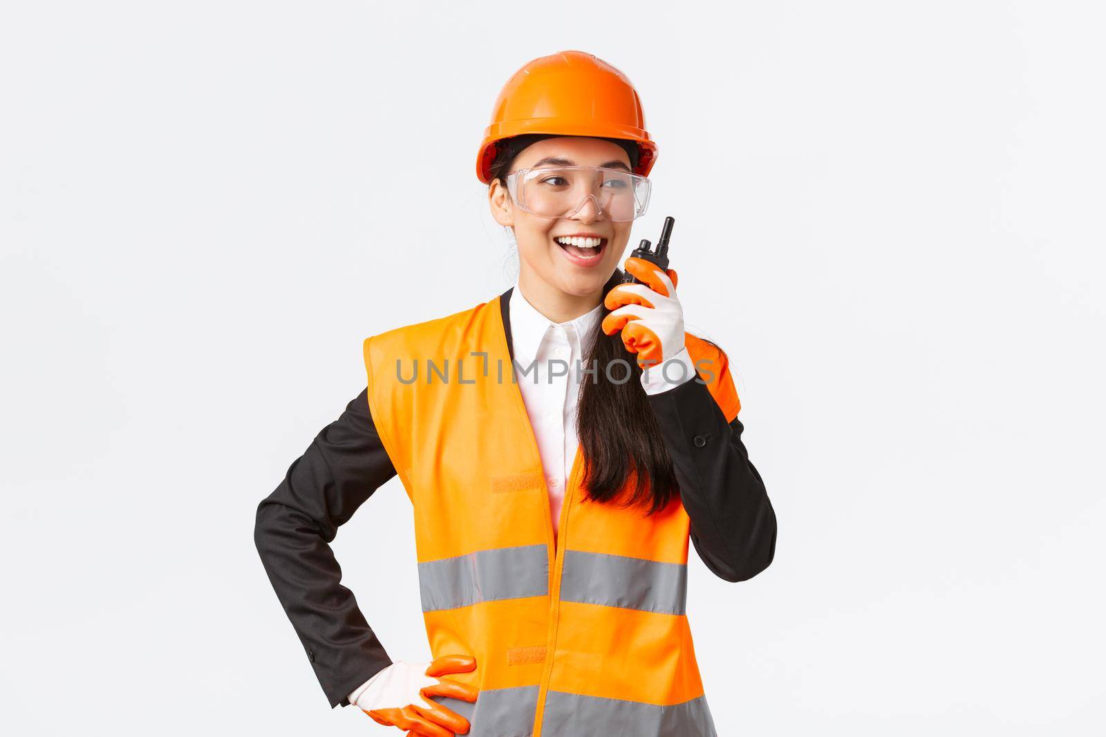 Confident pleased asian female engineer in safety helmet and uniform talking with chief architect using walkie-talkie. Satisfied construction technician contact team using radio phone by Benzoix