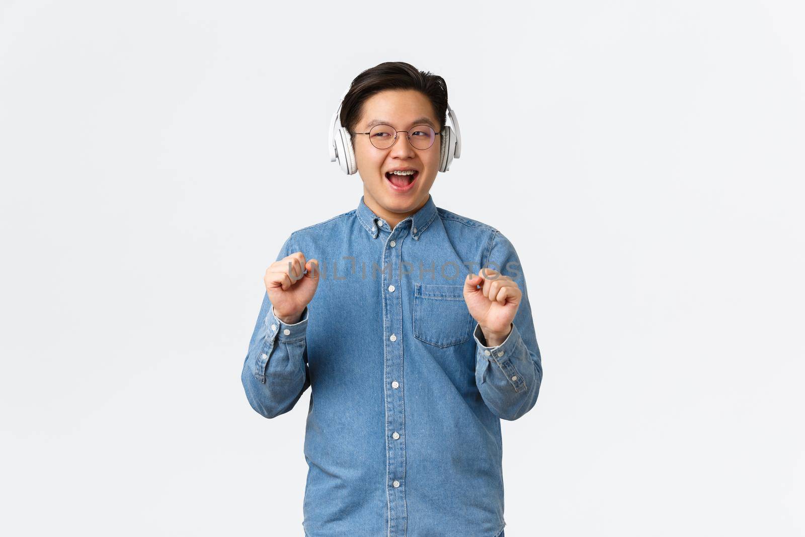 Lifestyle, leisure and technology concept. Carefree smiling funny asian guy vibing and chilling while listening music with wireless headphones, dancing and singing along, white background by Benzoix