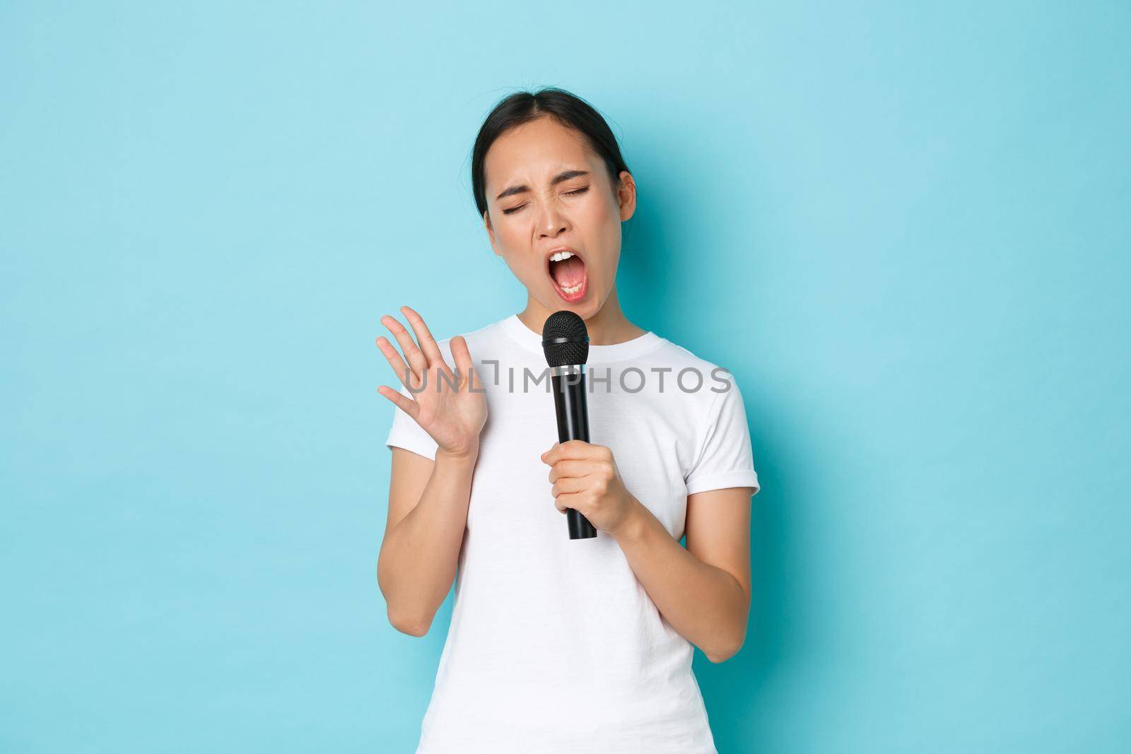 Lifestyle, people and leisure concept. Carefree beautiful asian girl singing sing in microphone with passionate expression, close eyes and gesturing while performing, like karaoke.