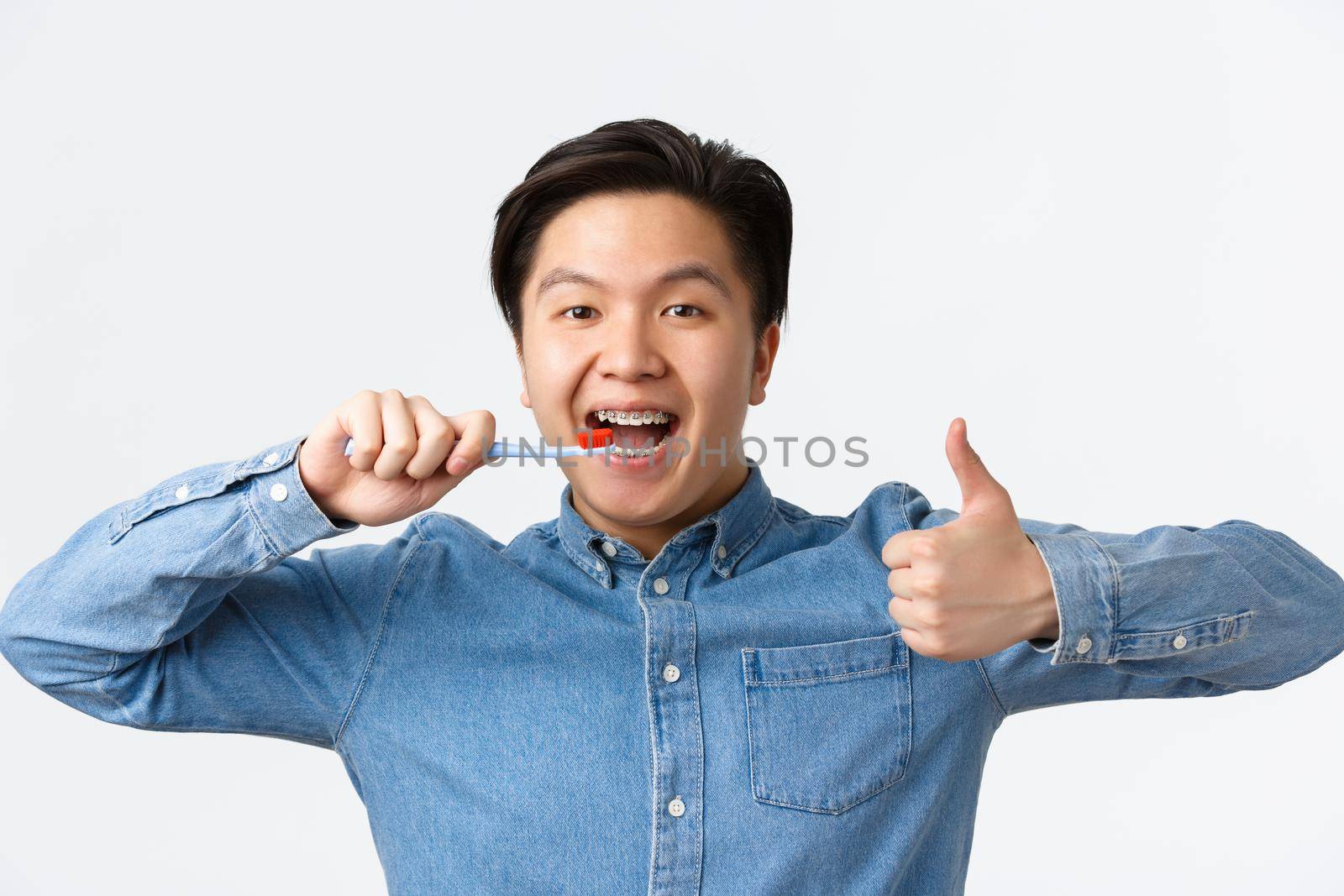 Orthodontics, dental care and hygiene concept. Close-up of satisfied happy asian man brushing teeth with braces, holding toothbrush and showing thumbs-up in approval, white background by Benzoix
