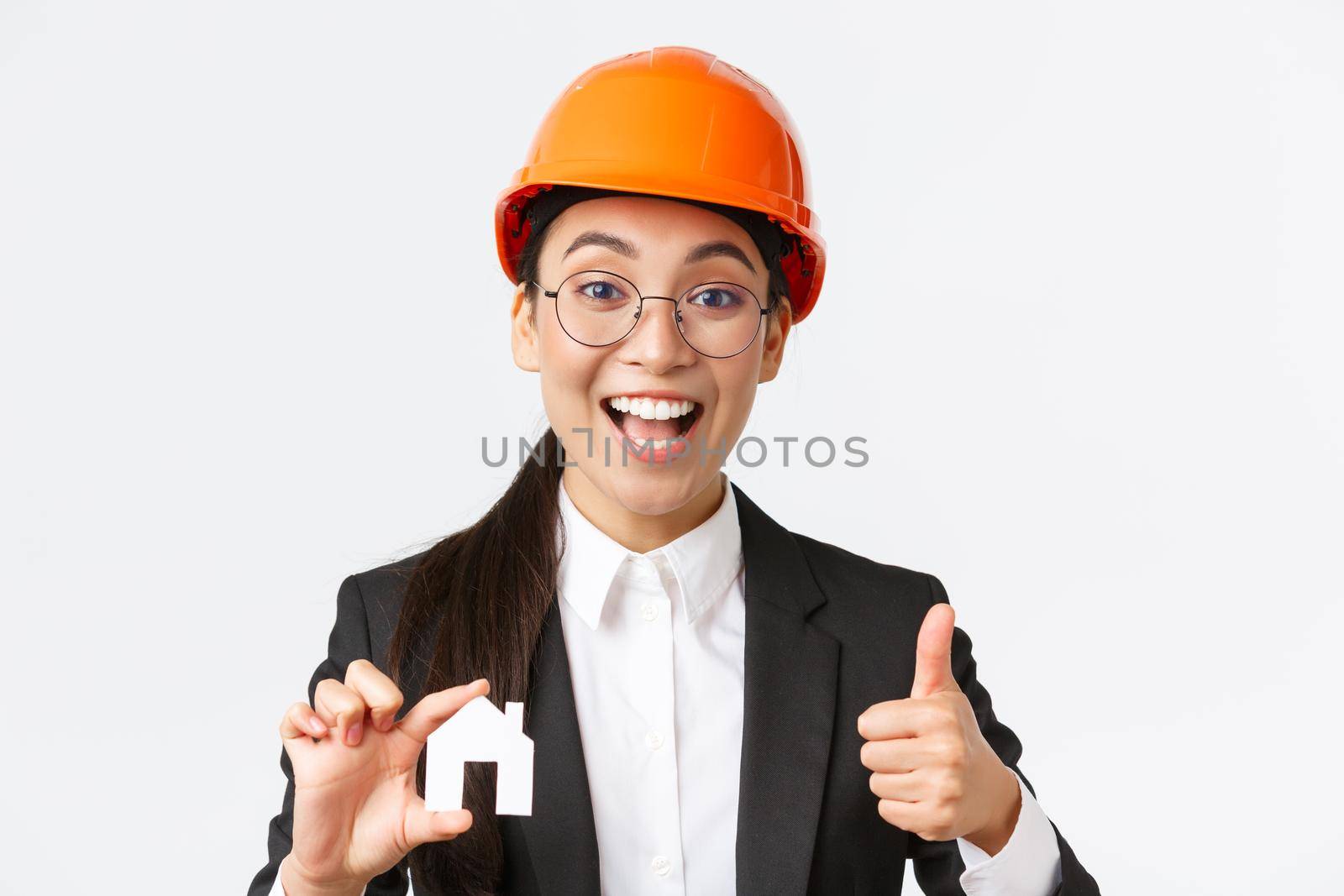 Close-up of upbeat professional female asian engineer, architect in helmet and business suit encourage contact her company for renovation and design, showing house and thumbs-up.