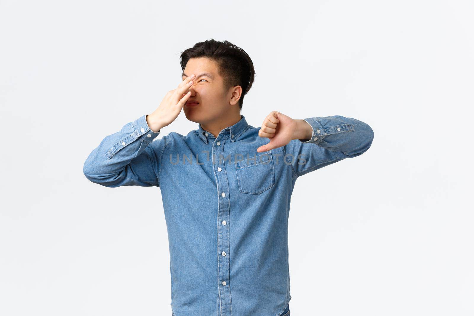 Disgusted asian man looking away and shut nose with fingers, showing thumbs-down, complaining awful reek, something stink, bothered with bad smell, standing white background.