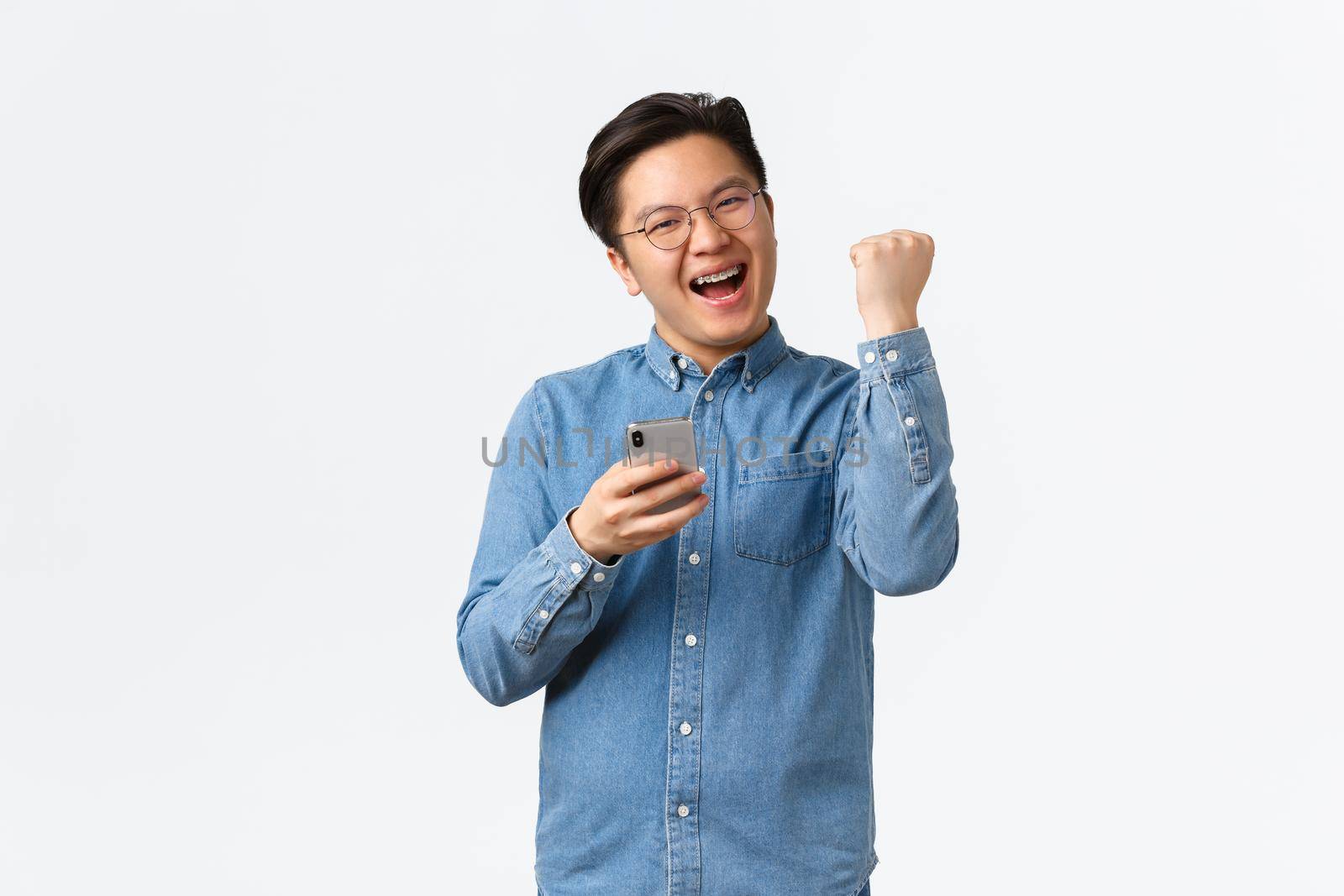 Satisfied, successful winning asian man in glasses and braces rejoicing, fist pump delighted and smiling as react to win in mobile game, receive positive feedback on application, white background by Benzoix