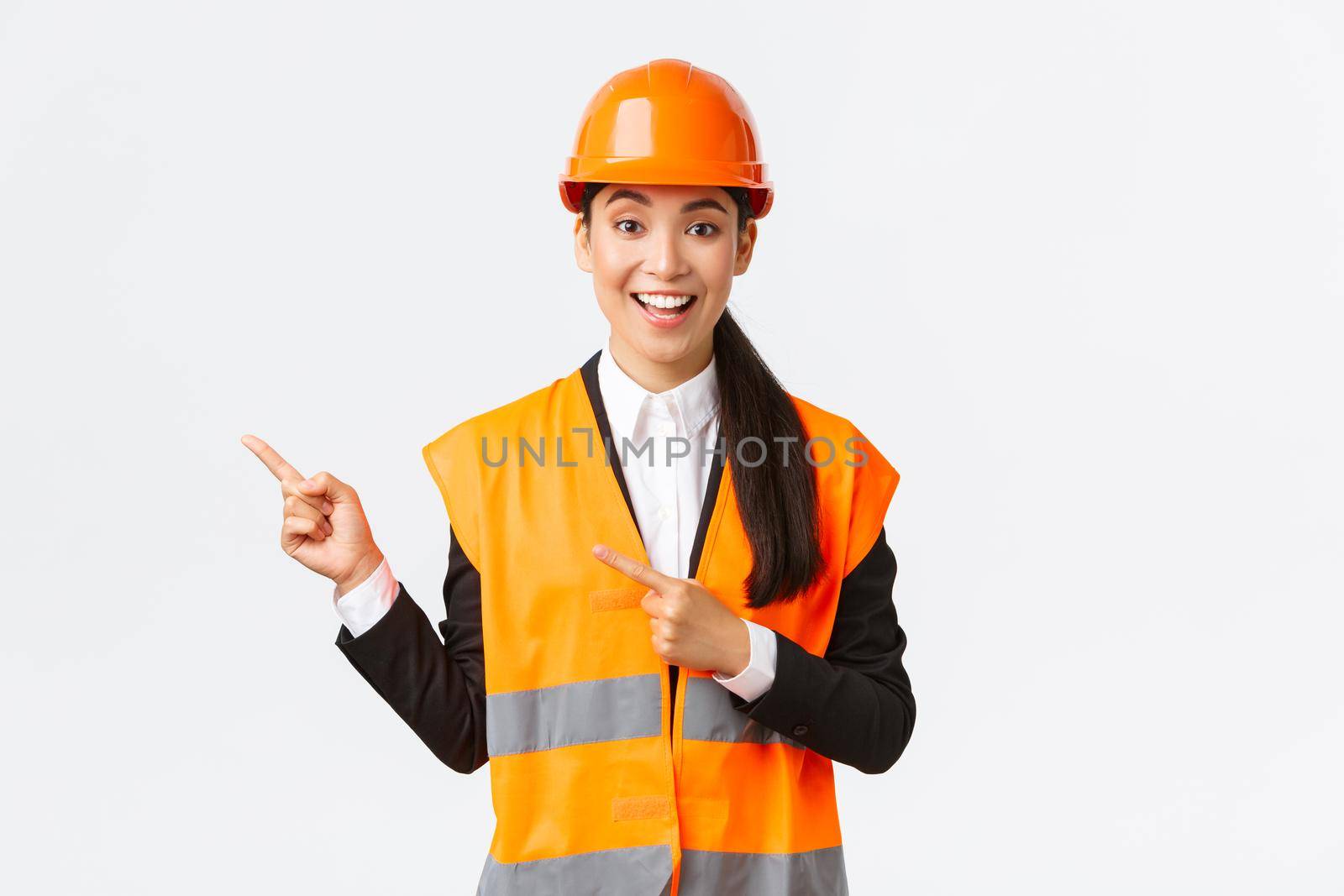 Building, construction and industrial concept. Smiling female engineer in helmet and reflective clothing showing way, pointing upper left corner, talking about constructing project, white background by Benzoix