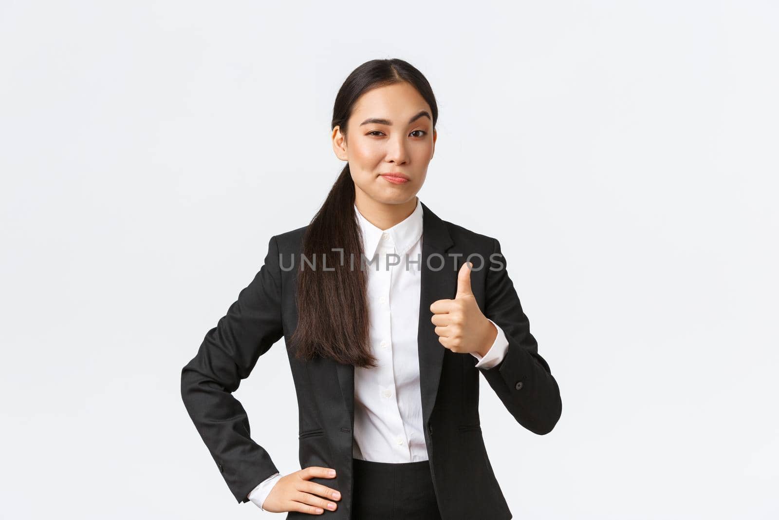 Pleased female entrepreneur, lady boss in black suit satisfied with your work, showing thumbs-up and nod in approval, praise great job, well done, standing white background by Benzoix