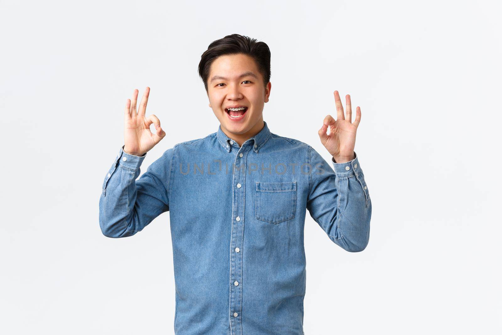 Smiling satisfied asian man with braces in blue shirt, showing okay gesture, congratulating person with excellent work, well done, recommend perfect service or quality, white background.