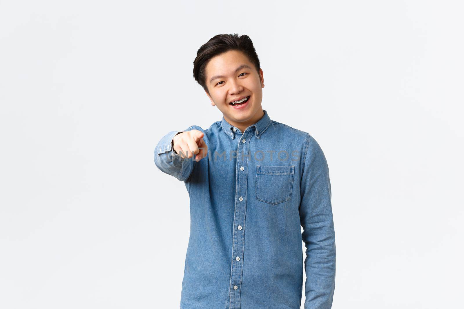 Smiling handsome asian guy with braces making choice, pointing finger at camera, choosing or picking something, found person, standing white background, congratulating you.