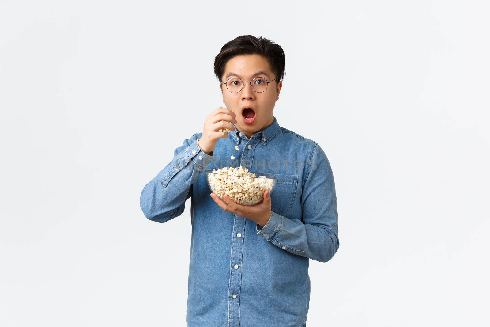 Leisure, lifestyle and people concept. Thrilled and amazed asian guy watching awesome movie or TV series, eating popcorn and looking astounished, standing white background.