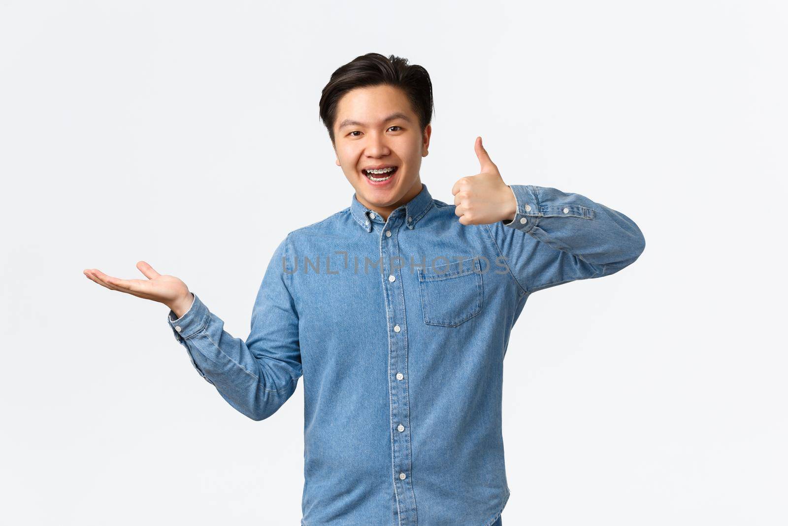 Satisfied handsome asian guy with braces in blue shirt, showing thumbs-up as rating product, raise hand over white background as if holding item, recommend it, demonstrate something by Benzoix