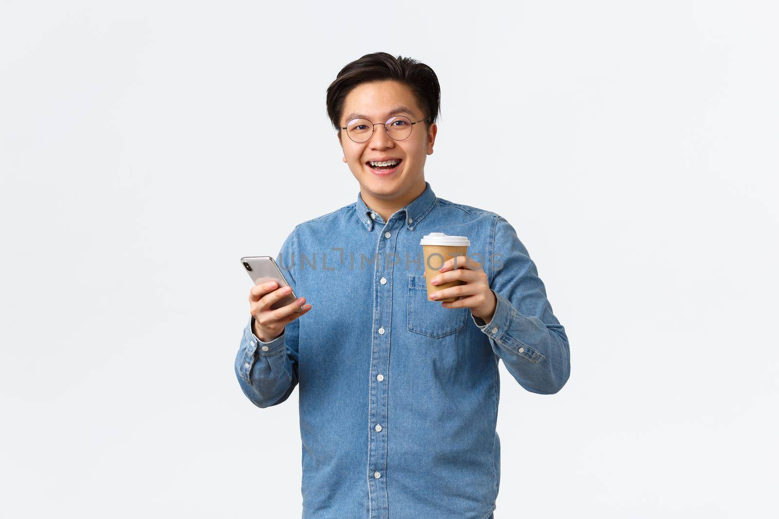 Lifestyle, leisure and technology concept. Carefree smiling asian man with braces, drinking coffee during lunch out of office, using mobile phone, texting friend, sending message, white background.